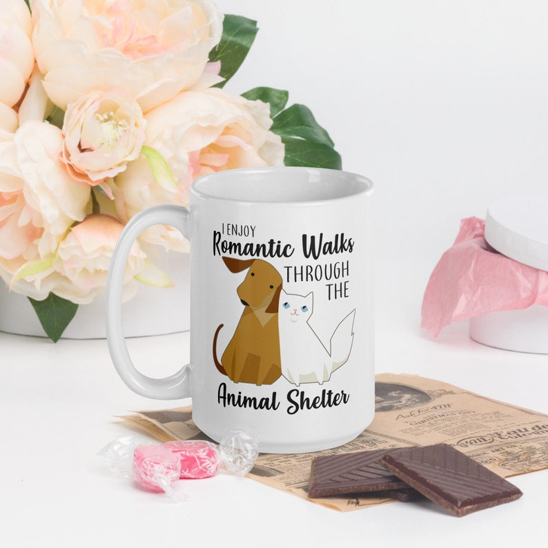 May include: White ceramic mug with a black and brown illustration of a dog and cat. The text on the mug reads "I enjoy romantic walks through the animal shelter."