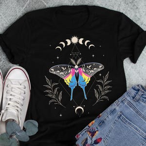 May include: Black t-shirt with a colorful butterfly design, moon phases, and stars. The butterfly is surrounded by a geometric pattern.