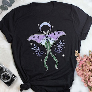 Può includere: T-shirt nero con un disegno di falena celeste. La falena ha ali lavanda con dettagli di luna crescente e stelle. Il corpo è verde e bianco, con lunghi viticci verdi. Rametti di foglie di lavanda e stelle circondano la falena.