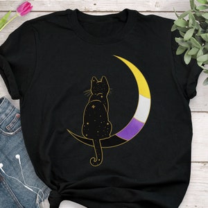 May include: Black t-shirt featuring a gold outline of a cat sitting on a crescent moon. The moon is colored with the non-binary pride flag. The cat's body is filled with stars.