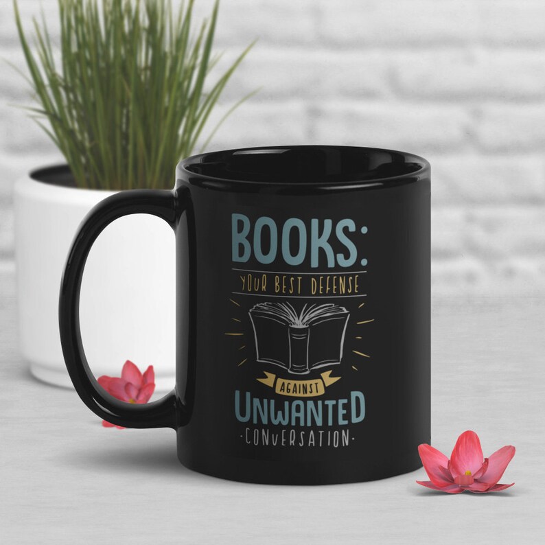 Puede incluir: Taza de cer&aacute;mica negra con el texto "BOOKS: YOUR BEST DEFENSE AGAINST UNWANTED CONVERSATION" en un dise&ntilde;o azul y blanco. La taza presenta una ilustraci&oacute;n de un libro abierto. Un regalo perfecto para los amantes de los libros.