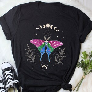 Op de afbeelding: Zwart T-shirt met een kleurrijk vlinderontwerp, maanfasen en geometrische vormen. De vlinder heeft roze, groene en blauwe vleugels.