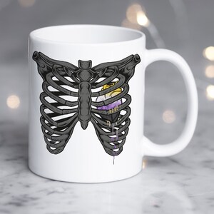Può includere: Tazza in ceramica bianca con un'illustrazione in bianco e nero di una gabbia toracica con un cuore viola, giallo e bianco al suo interno.