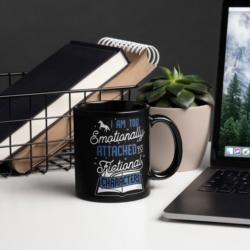 May include: Black coffee mug with a white design that says "I am too emotionally attached to fictional characters". The mug is sitting on a white table next to a laptop and a potted plant.