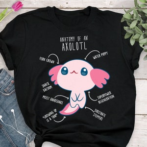 May include: Black t-shirt featuring a pink cartoon axolotl and white text that reads "Anatomy of an Axolotl" with arrows pointing to different parts of the axolotl and labels such as "Fern Crown", "Water Puppy", "Snack Vacuum", "Moist Handshake", "Superpower: Regeneration", "Substrate Steppers", and "Growing Up is a Trap".
