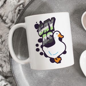 May include: White ceramic mug with a black and green graphic of a goose and the text "Gay As" in a comic book style font.