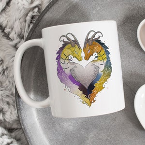 May include: White ceramic mug featuring a heart-shaped design of two dragons. The dragons are yellow and orange, with rainbow-colored wings and tails. A gray heart is in the center of the dragons. The mug has a white handle.