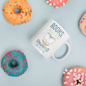 Puede incluir: Taza de cer&aacute;mica blanca con el texto "BOOKS: YOUR BEST DEFENSE AGAINST UNWANTED CONVERSATION" y una ilustraci&oacute;n de un libro abierto. La taza est&aacute; rodeada de coloridos donuts con chispas y glaseado sobre una superficie azul claro.