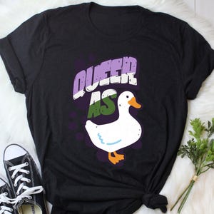 May include: Black t-shirt with the text "Queer as" in purple, green, and white, and a white goose with orange feet.
