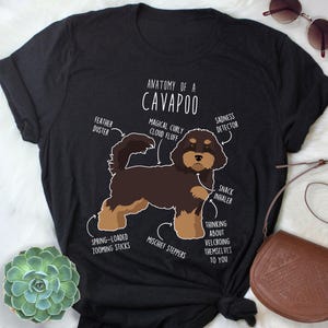 May include: Black t-shirt featuring a cartoon illustration of a Cavapoo dog with labeled body parts. The labels include "Feather Duster," "Magical Curly Cloud Fluff," and "Sadness Detector." The shirt design is humorous and dog-themed.