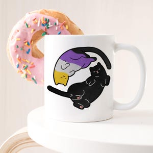 Op de afbeelding: Witte keramische mok met een zwart en paars kattenontwerp. De kat is opgerold in een yin-yang symbool met de kleuren van de non-binaire pride-vlag.