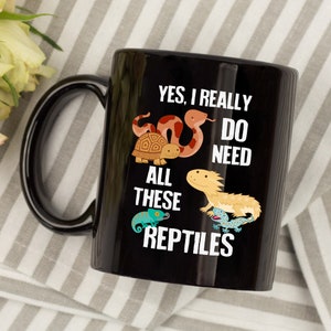 K&ouml;nnte beinhalten: Schwarze Keramik-Kaffeetasse mit einem wei&szlig;en Design, das eine Schlange, eine Schildkr&ouml;te, einen Bartagamen und einen Cham&auml;leon zeigt. Der Text lautet "Ja, ich brauche wirklich all diese Reptilien".