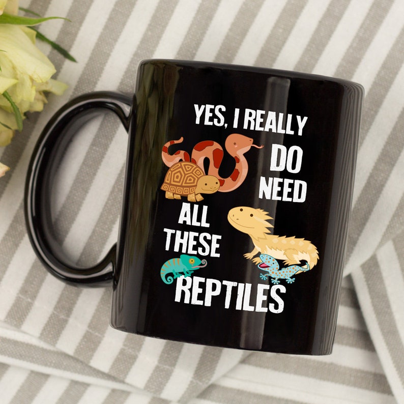 K&ouml;nnte beinhalten: Schwarze Keramik-Kaffeetasse mit einem wei&szlig;en Design, das eine Schlange, eine Schildkr&ouml;te, einen Bartagamen und einen Cham&auml;leon zeigt. Der Text lautet "Ja, ich brauche wirklich all diese Reptilien".