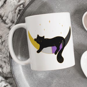 Puede incluir: Taza de cerámica blanca con un gato negro durmiendo en una luna creciente. La luna está decorada con los colores de la bandera asexual: negro, blanco, morado y gris. Hay estrellas doradas en el fondo.