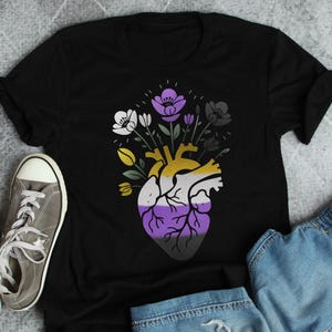 May include: A black short-sleeve t-shirt featuring a graphic of an anatomical heart with a color scheme of yellow, white, purple, and black. The heart is topped with a bouquet of flowers in matching colors, including white, purple, and black petals.