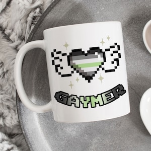 Może przedstawiać: Biały kubek ceramiczny z pikselowanym sercem w kolorze zielonym, szarym i czarnym z skrzydłami i napisem "Gaymer" na banerze poniżej.