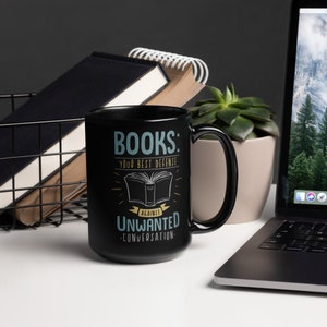 Puede incluir: Taza de cer&aacute;mica negra con el texto "BOOKS: your best defense against unwanted conversation" en verde azulado y blanco. La taza presenta una ilustraci&oacute;n de un libro abierto. Otros elementos en la imagen incluyen un port&aacute;til, libros y una planta suculenta.