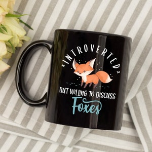 May include: Black coffee mug with a white graphic of a cartoon fox and the text "Introverted but willing to discuss foxes".