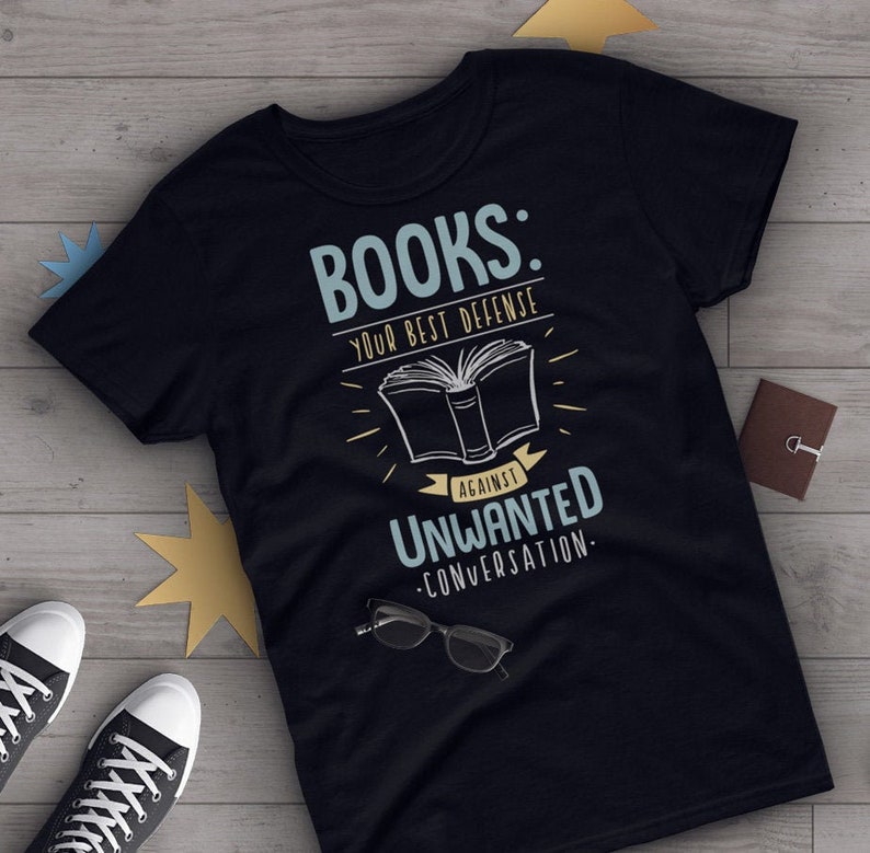 以下が含まれることがあります： 黒いTシャツに、白い文字で「本：不要な会話に対する最高の防御」と書かれています。グラフィックには、開いた本のスタイリッシュな画像が含まれています。