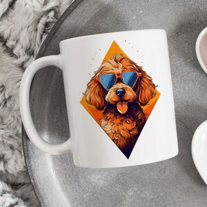 Puede incluir: Taza de cerámica blanca con un caniche marrón que lleva gafas de sol naranjas en forma de diamante amarillo. El caniche tiene la lengua fuera.