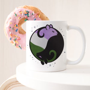 May include: White ceramic mug featuring a non-binary themed design with a yin-yang style illustration of two stylized animals in purple, black, and green. A pink frosted donut with sprinkles is next to the mug.