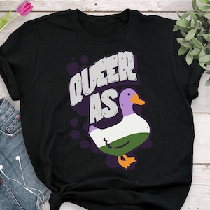 May include: Black t-shirt with the text "Queer as" and a cartoon duck with purple, white, and green feathers.