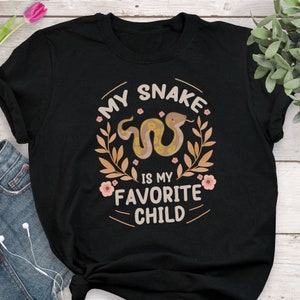 Puede incluir: Camiseta negra con el texto "MY SNAKE IS MY FAVORITE CHILD" en un diseño circular. El diseño incluye una serpiente de dibujos animados, hojas y flores en tonos marrón, beige y rosa.