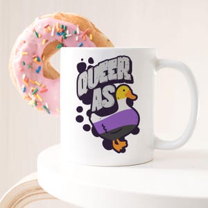 May include: White ceramic mug with a black and white graphic of a duck with a purple, black, and white striped body. The text "Queer as" is above the duck.
