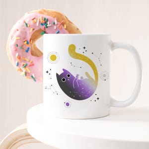 Puede incluir: Taza de cerámica blanca con un gato negro con una cola degradada en morado y amarillo. El gato está rodeado de estrellas y planetas. La taza está sobre una superficie blanca.