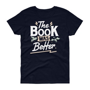 Pode incluir: Uma camiseta azul marinho com texto branco que diz "The Book Was Better" com um raio estilizado antes e depois de "The" e um raio estilizado abaixo de "Better". H&aacute; um unic&oacute;rnio branco e um drag&atilde;o verde de cada lado do texto.