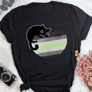 Può includere: T-shirt nero con un gatto nero dei cartoni animati su un motivo a forma di cuore. Il cuore presenta strisce orizzontali grigie, bianche e verdi. Il gatto ha occhi bianchi e un contorno bianco.