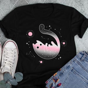 Peut inclure: T-shirt noir avec un motif de chat galaxie rose et noir. Le chat est enroulé en boule avec sa queue enroulée autour de son corps. Le motif est entouré d'étoiles et de planètes roses.