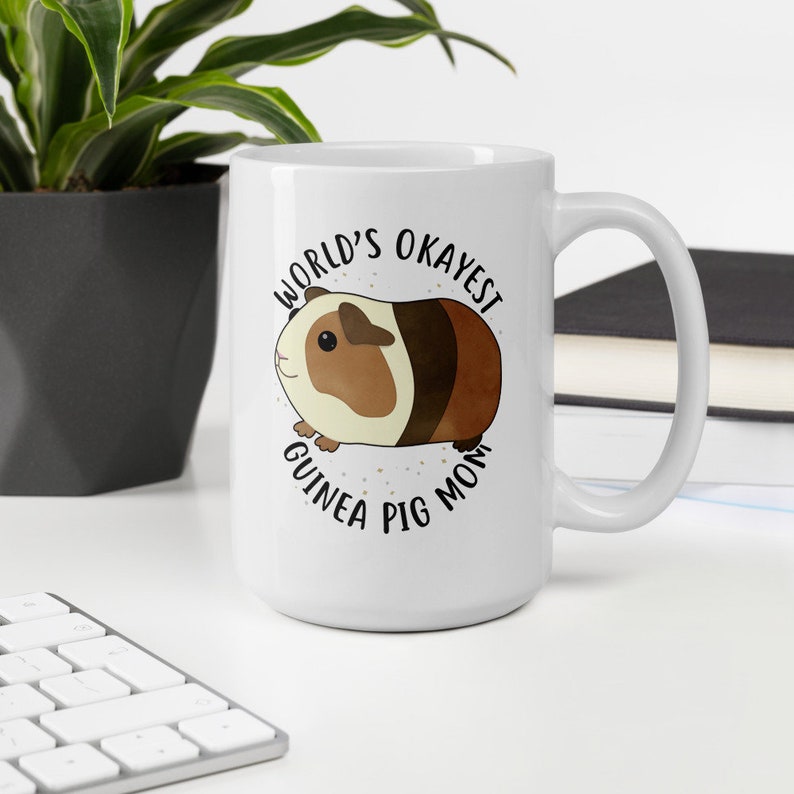 May include: White ceramic mug with a brown and white guinea pig illustration and the text "World's Okayest Guinea Pig Mom".