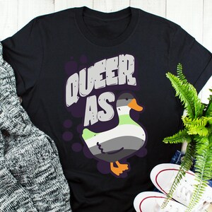 May include: Black t-shirt with the text "Queer as" and a cartoon duck with a green, gray, and white color scheme.