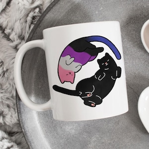 Può includere: Tazza in ceramica bianca con un design di gatto nero e viola. I gatti sono arrotolati in un cerchio, con il gatto viola sopra il gatto nero. Il design è un simbolo della bandiera del pride asessuale.
