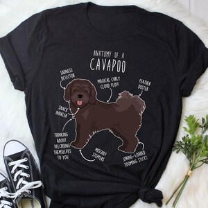 May include: A black t-shirt with a white graphic of a brown Cavapoo dog. The graphic includes text that says "Anatomy of a Cavapoo" and labels different parts of the dog, such as "Magical Curly Cloud Fluff", "Snack Inhaler", and "Spring-Loaded Zooming Sticks".