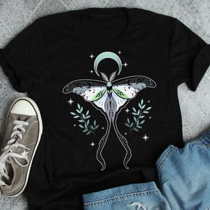 Może przedstawiać: Czarny t-shirt z motywem ćmy, półksiężycem i gwiazdami. Ćma ma szare, białe i zielone skrzydła ze skomplikowanymi wzorami. Ozdobne gałązki i gwiazdy otaczają ćmę, tworząc niebiański motyw.