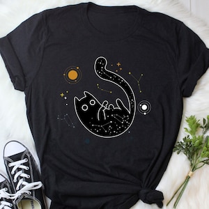 May include: Black t-shirt featuring a graphic of a black cat with white stars inside its silhouette, floating in space with a planet and constellations.