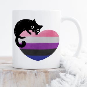 Puede incluir: Taza de cerámica blanca con una ilustración de un gato negro sobre un diseño en forma de corazón. El corazón muestra los colores de la bandera del orgullo genderqueer: rosa, blanco, gris, morado, negro y azul. La taza tiene un asa curva.