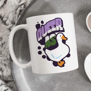 May include: White ceramic mug with a graphic of a white duck and the text "Queer as" in purple, green, and white colors.