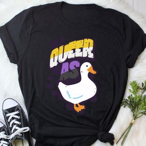 May include: Black t-shirt with the text "Queer as" in yellow, purple, and black lettering. A white duck with a yellow beak and feet is below the text.
