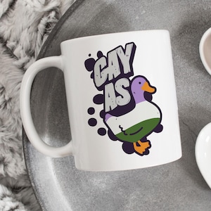 May include: White ceramic mug with the text "GAY AS" in gray letters. A cartoon duck with a purple head, white body, green bottom, and orange feet is featured on the mug. Black ink splatters surround the design.