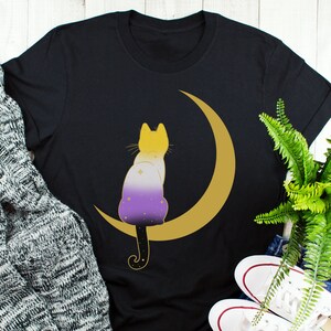 May include: Black t-shirt with a yellow crescent moon and a purple, white, and yellow cat sitting on the moon. The cat has stars on its body.