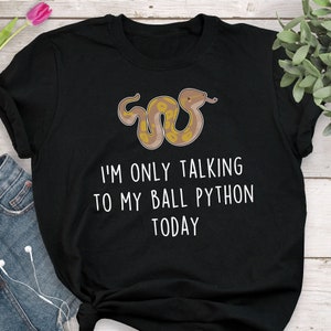 May include: Black t-shirt featuring a cartoon ball python with a yellow and brown pattern. The text "I'M ONLY TALKING TO MY BALL PYTHON TODAY" is printed in white below the snake illustration. The shirt is made of a soft material.