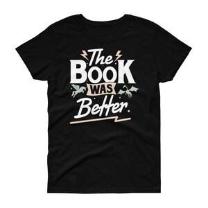 Pode incluir: Camiseta preta com texto branco que diz "The Book Was Better" com um raio antes e depois de "The" e um raio abaixo de "Better". H&aacute; duas pequenas ilustra&ccedil;&otilde;es de criaturas m&iacute;ticas, um unic&oacute;rnio e um drag&atilde;o, de cada lado do texto.