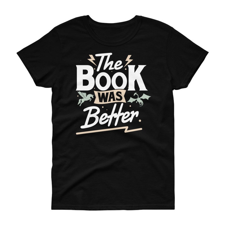Pode incluir: Camiseta preta com texto branco que diz "The Book Was Better" com um raio antes e depois de "The" e um raio abaixo de "Better". H&aacute; duas pequenas ilustra&ccedil;&otilde;es de criaturas m&iacute;ticas, um unic&oacute;rnio e um drag&atilde;o, de cada lado do texto.