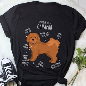 May include: A black t-shirt with a white graphic of a brown Cavapoo dog. The graphic includes text that says "Anatomy of a Cavapoo" and lists different features of the dog, such as "Magical Curly Cloud Fluff", "Feather Duster", "Snack Inhaler", and "Spring-Loaded Zooming Sticks".