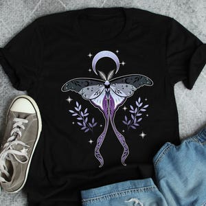 以下が含まれることがあります： 黒いTシャツ。蛾のデザイン、三日月、星のアクセントが特徴です。蛾はグレー、紫、白のディテールです。デザインにはラベンダー色の葉の小枝が含まれています。グレーのスニーカーとデニムショーツが見えます。