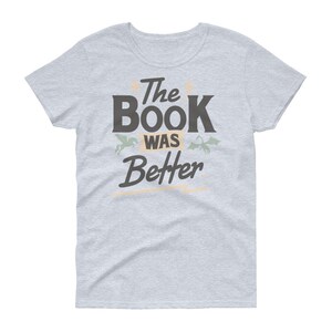 Pode incluir: Uma camiseta cinza claro com o texto "The Book Was Better" em preto. O texto &eacute; estilizado com uma fonte vintage e &eacute; cercado por duas pequenas ilustra&ccedil;&otilde;es, uma de um drag&atilde;o e outra de um unic&oacute;rnio.