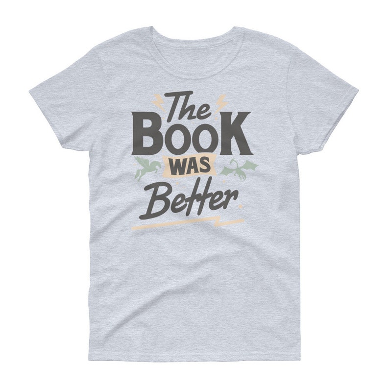 Pode incluir: Uma camiseta cinza claro com o texto "The Book Was Better" em preto. O texto &eacute; estilizado com uma fonte vintage e &eacute; cercado por duas pequenas ilustra&ccedil;&otilde;es, uma de um drag&atilde;o e outra de um unic&oacute;rnio.